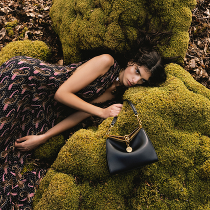 girl-lying-in-a-forest-in-a-flounced-dress-with-micro-Pailsey-holds-a-black-Vela-bag-in-her-hands.