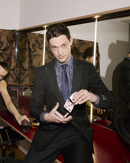 A man wearing a tailored black brocade suit by ETRO, paired with a paisley-patterned tie, performs a card trick in a refined backstage environment.