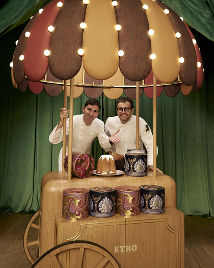 Two chefs in white uniforms stand smiling behind a wooden dessert cart decorated with colorful paisley-patterned tins and an umbrella canopy with warm lights. The cart has a rustic design with "ETRO" engraved on the front, and the display features a pandoro or panettone cake. A green curtain backdrop adds a festive atmosphere.