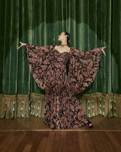 A woman poses gracefully in an elaborate, flowing gown with a paisley-inspired pattern in rich tones of pink, black, and purple. The dress features dramatic, wide sleeves that she extends outward, creating a wing-like effect. She stands on a wooden stage in front of a green velvet curtain with decorative fringe, exuding elegance and theatrical flair.