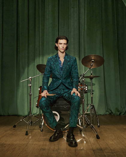 A person in a teal patterned suit sits on a drum kit stool in front of drums, against green curtains.