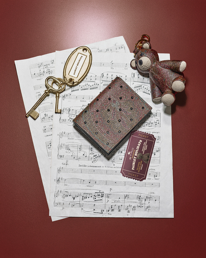 A photograph showing a stylish arrangement on a red surface. The composition includes: Sheets of classical music notations, featuring the title "Tristan und Isolde", A pair of gold keys attached to a keychain with an oval tag engraved with "Palais Holiday”, A textured, small wallet or cardholder with an intricate pattern and decorative accents, A teddy bear keychain in a multicolored fabric, A vintage-style ticket labeled "Palais Holiday," completing the aesthetic ensemble.