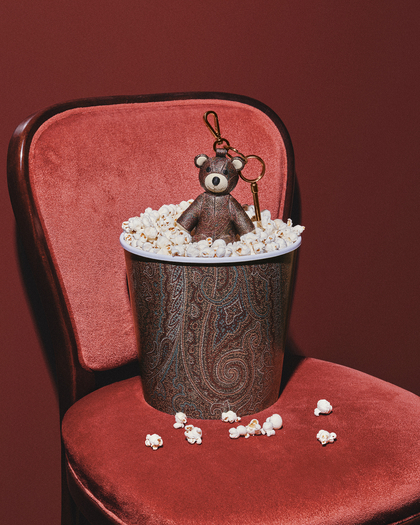 A cute teddy bear keychain sitting atop a cup filled with popcorn, featuring a decorative patterned design on the cup and a warm red background.