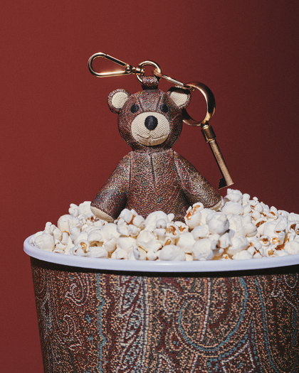 A cute teddy bear keychain sitting atop a cup filled with popcorn, featuring a decorative patterned design on the cup and a warm red background.