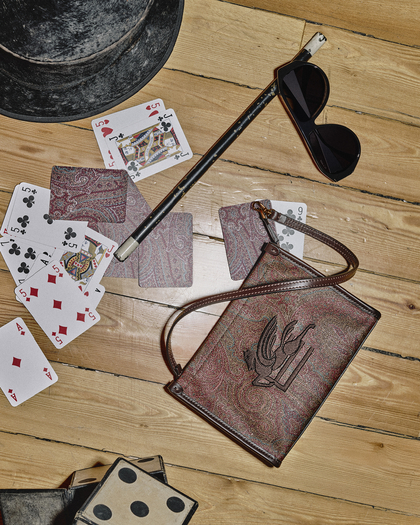  A flat-lay composition featuring a vintage-inspired scene on a wooden floor. A distressed black top hat sits in the corner, surrounded by playing cards, including face cards and numbered cards, scattered across the surface. A slender black wand with white tips lies diagonally near a pair of angular black sunglasses. A small, paisley-patterned bag with a bird emblem and a thin leather strap is placed prominently, with its earthy tones complementing the wooden floor. Nearby, a pair of oversized dice adds a playful, magical theme to the arrangement.