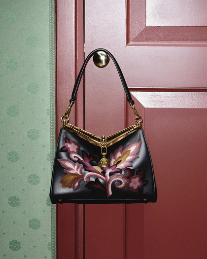 A photograph of an Embroidered Vela bag hanging on the brass knob of a closed, deep red door. The bag is crafted in sleek black leather and adorned with intricate floral embroidery in shades of pink, purple, and gold, adding a bold yet sophisticated touch. It features a striking gold metal frame and hardware, including a golden medallion charm. The black leather strap is attached with polished gold accents. The backdrop includes a green patterned wall, creating a stylish contrast against the vivid red door and the bag’s luxurious design.