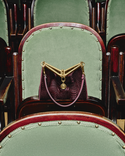 A photograph of a Velvet Vela bag placed on a green velvet theater seat with polished wooden edges, studded with metallic rivets. The handbag features a striking design with a deep burgundy fabric patterned in wavy ridges, complemented by a golden metallic frame and hardware. A pink strap adds a contrasting touch, and a decorative golden medallion charm hangs from the bag, enhancing its elegant appearance. The scene suggests a sophisticated and vintage setting, likely an opera house or classical theater.