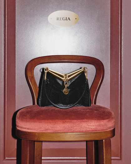 A photograph of a sophisticated black Velvet Vela bag placed on a wooden chair with a plush, rust-red velvet seat. The handbag features a textured wavy pattern, a gold metallic frame, and a decorative medallion charm. Its elegant design is further enhanced by the black strap attached with polished gold hardware. The background includes a deep red wall with a plaque reading "REGIA," emphasizing a luxurious and refined setting. The combination of the chair, the handbag, and the decor creates a regal and stylish atmosphere.