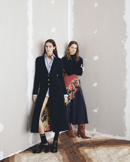 Two women posing in a minimalist room with a rug and partially unfinished walls. The woman on the left is wearing a long, black coat with gold buttons, a light blue shirt, and black platform boots, revealing a floral-patterned skirt through the open coat. The woman on the right is dressed in a black sweater and a long, dark skirt with a floral design, paired with brown leather boots.