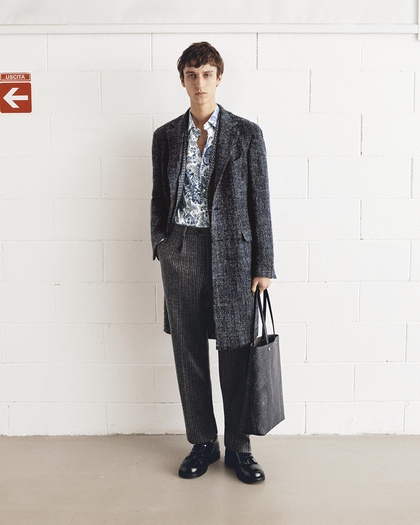 A young man standing against a white brick wall, wearing a dark textured overcoat over a patterned blue shirt, and gray pinstripe trousers. He is holding a black leather tote bag in one hand and is wearing black polished shoes. The overall look is stylish and modern, with a neutral expression on his face.