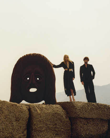 FW24 ADV fashion campaign - A man and a woman standing on large stone blocks outdoors, with a mountainous landscape in the background. The woman, wearing a dark outfit with a midriff cutout, rests her hand on a large, ominous, dark brown mask sculpture with glowing white eyes and an open mouth. The man, dressed in a black suit, stands slightly behind her, both gazing towards the camera in a surreal, eerie setting.