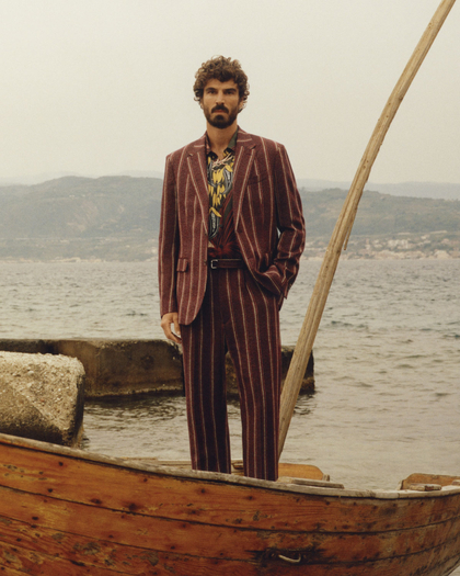 A man with curly hair and a beard standing on a wooden rowboat, wearing a striped maroon suit over a patterned shirt. The boat is in shallow water near large rocks, with a backdrop of calm water and distant mountains under an overcast sky