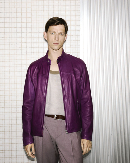 The model wears a purple leather jacket over a striped tank, paired with taupe trousers and a brown belt