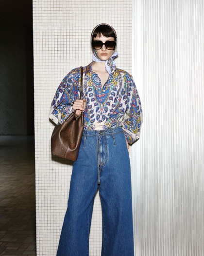 A fashionable woman wearing a colorful patterned blouse, wide-leg jeans, a headscarf, and oversized sunglasses, carrying a brown leather bag.