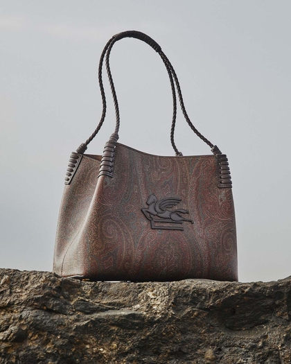 The image shows an elegant brown leather Libra bag with two long braided handles. The surface of the bag features an intricate paisley pattern, with warm tones of red and brown. At the center, there is a large embossed design of a winged horse, adding a bold and distinctive element to the otherwise subtle texture of the bag. The bag is placed on a rugged stone surface, with the sky as the backdrop, highlighting its refined craftsmanship in contrast to the natural setting.