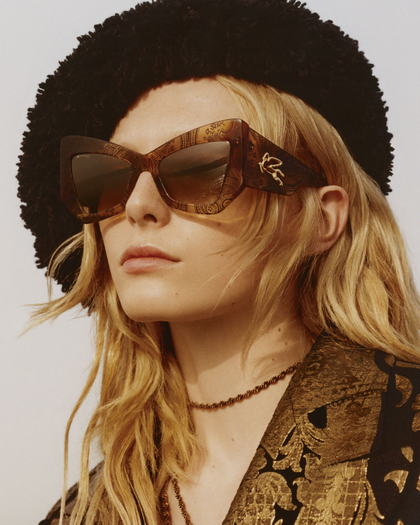 A model with long blonde hair wears large, decorative sunglasses and a black beret-style hat. The outfit is complemented by a richly patterned jacket and a thin necklace. The overall look conveys a vintage and sophisticated style.