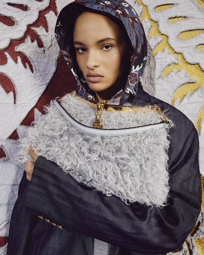 A model with a serious expression wearing a dark, patterned hooded jacket, holding a fluffy, gray textured Vela bag with gold accents against a backdrop of intricately designed fabric featuring white, red, and yellow patterns