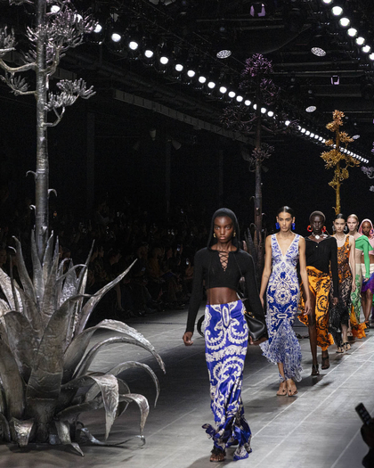 MAKING ETRO, SS25 woman collection: The image shows a fashion runway with models walking in a line, wearing vibrant and intricately designed outfits in various colors, including blue, orange, green, and black. The designs appear to feature bold patterns and textures. The runway is flanked by large metallic sculptures resembling plants, with one particularly prominent silver sculpture in the foreground. The lighting above illuminates the models as they walk down the catwalk, while the audience on either side watches. The overall atmosphere is striking and artistic, with an emphasis on high fashion and creative set design