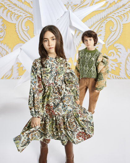 Two children posing against a decorative, ornate background featuring a large paper origami design and intricate yellow and white patterns. The child in the foreground has long, dark hair and is wearing a loose, flowing paisley dress with earthy tones of green, red, and blue, paired with brown boots. The child in the background has shorter, wavy dark hair and is dressed in a green and beige knit sweater with a textured design, brown pants, and a serious expression. The overall atmosphere is artistic and fashion-forward.