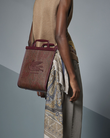 Person holding a burgundy tote with an embossed winged lion, wearing a tan top, gray pants, and a patterned scarf.