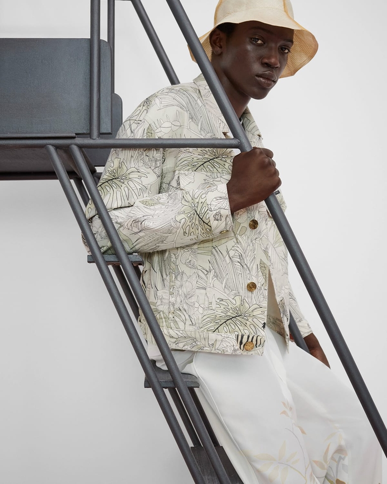 Homme sur les escaliers avec chapeau forme Bob qui porte une veste en jean imprimé tropical et zèbres et jean blanc 