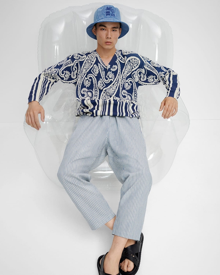 Young man sitting on an inflatable chair wearing paisley denim bucket hat with logo, striped paisley cotton jumper- SS23 man collection