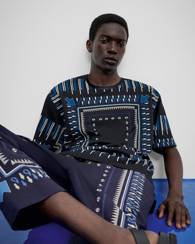 Sitzender junger Mann mit Baumwollhemd mit Bandana-Print, Jogginghose und Ledersandalen - Herrenkollektion FS23