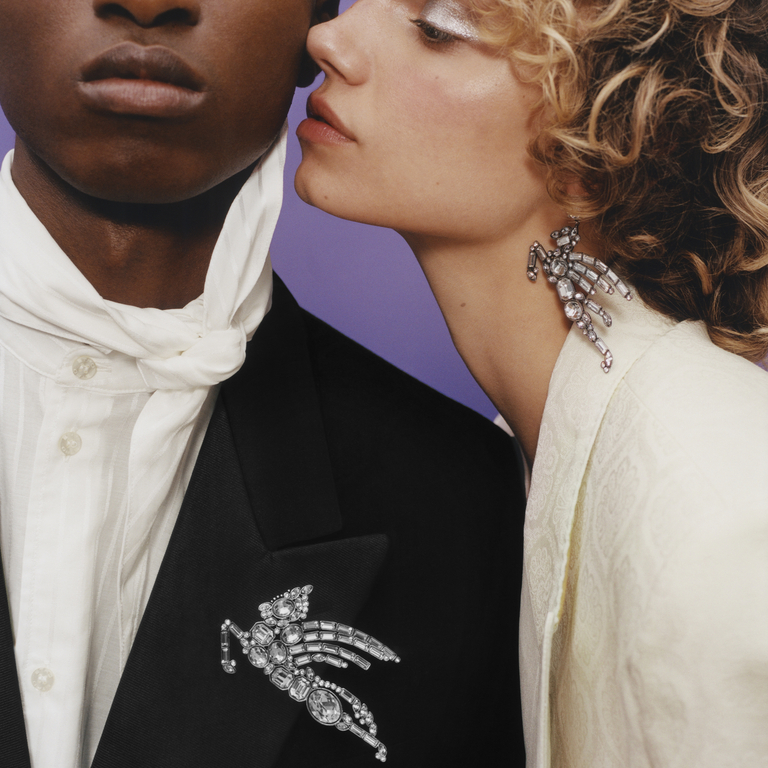 Two models in the foreground are wearing ETRO PE24 shirts and jackets. He wears a maxi Pegaso brooch with rhinestones on the lapel of his jacket. She wears a maxi Pegaso earring with rhinestones